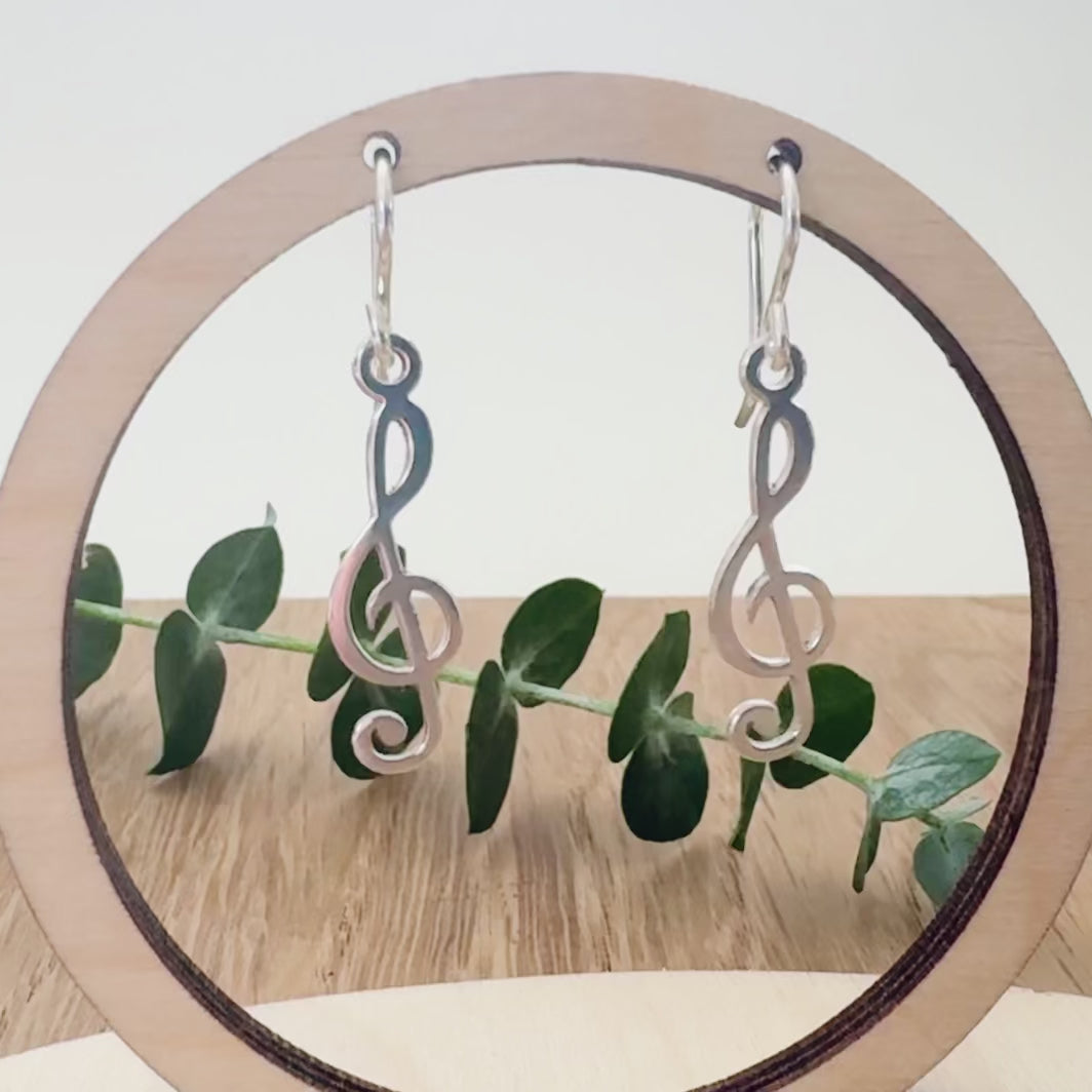 Sterling silver earrings with a treble clef design on a wooden surface with a small sprig of eucalyptus leaves.