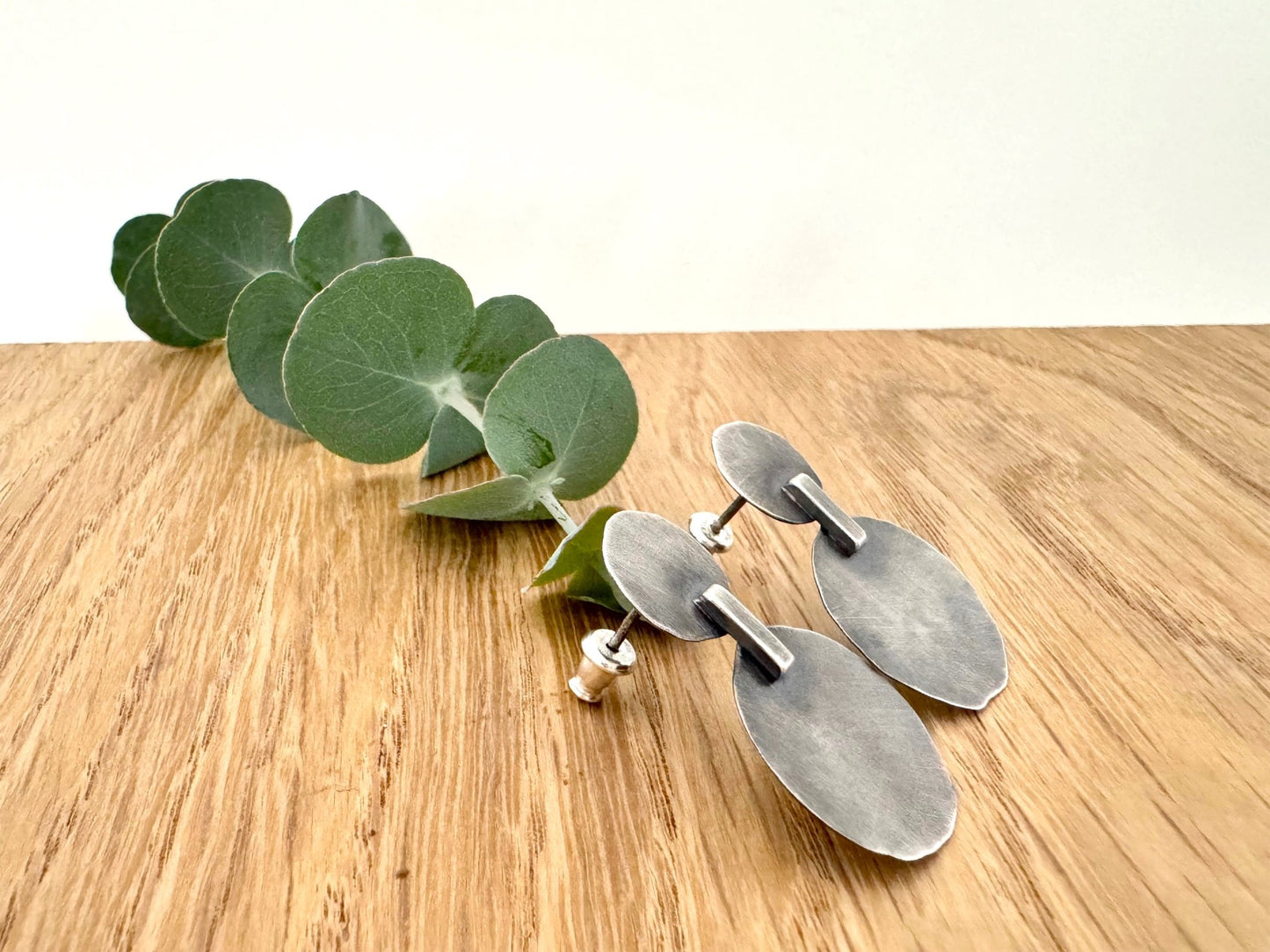 Pair of silver earrings on a wooden surface with a sprig of eucalyptus leaves.