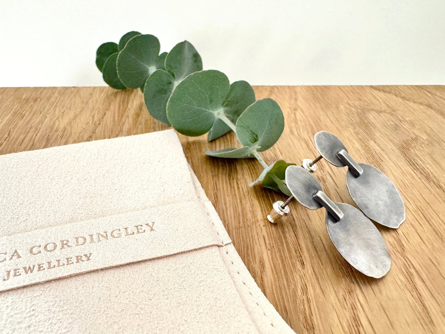 Silver earrings on a wooden surface with a pouch labelled 'Rebecca Cordingley Jewellery'.