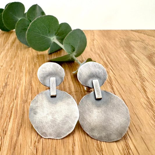 Pair of silver earrings on a wooden surface with green leaves in the background