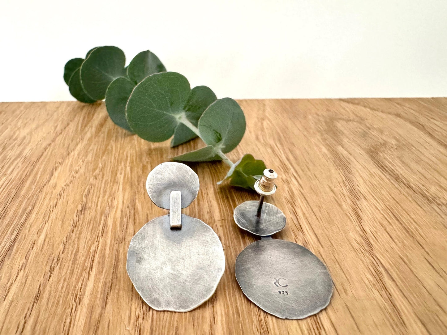 Silver earrings on a wooden surface with a small sprig of eucalyptus leaves