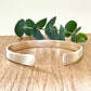 A sterling silver cuff bracelet with flared ends and a brush finish, on a wooden background with a small sprig of eucalyptus leaves.