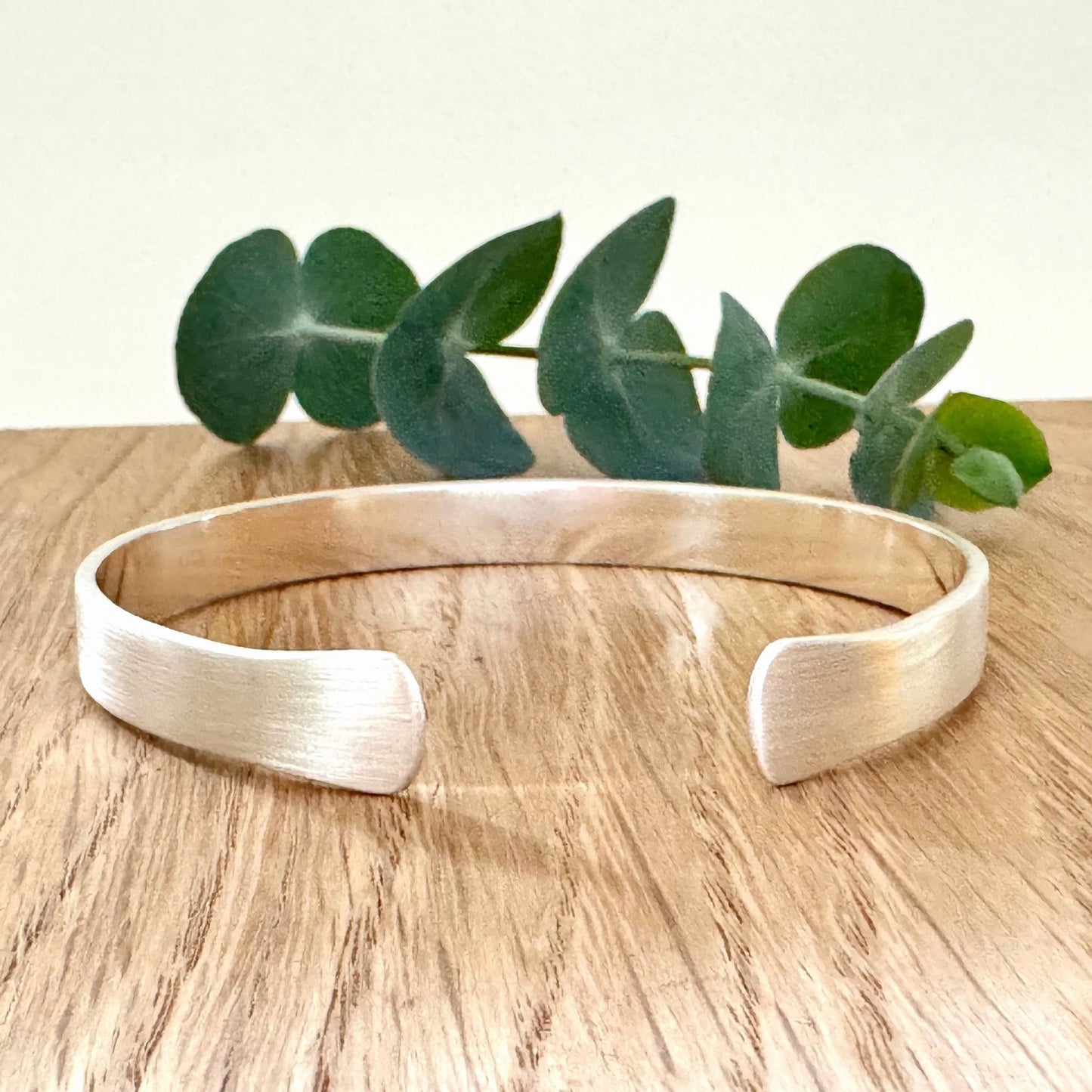 A sterling silver cuff bracelet with flared ends and a brush finish, on a wooden background with a small sprig of eucalyptus leaves.