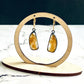 A pair of handmade sterling silver drop earrings featuring gold rutile quartz gemstones. The sterling silver bezel settings have been heavily oxidised and the earwires are 14k gold fill lever backs. The earrings are hanging from a wooden stand, which is on a white marble base with a black background.