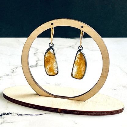 A pair of handmade sterling silver drop earrings featuring gold rutile quartz gemstones. The sterling silver bezel settings have been heavily oxidised and the earwires are 14k gold fill lever backs. The earrings are hanging from a wooden stand, which is on a white marble base with a black background.