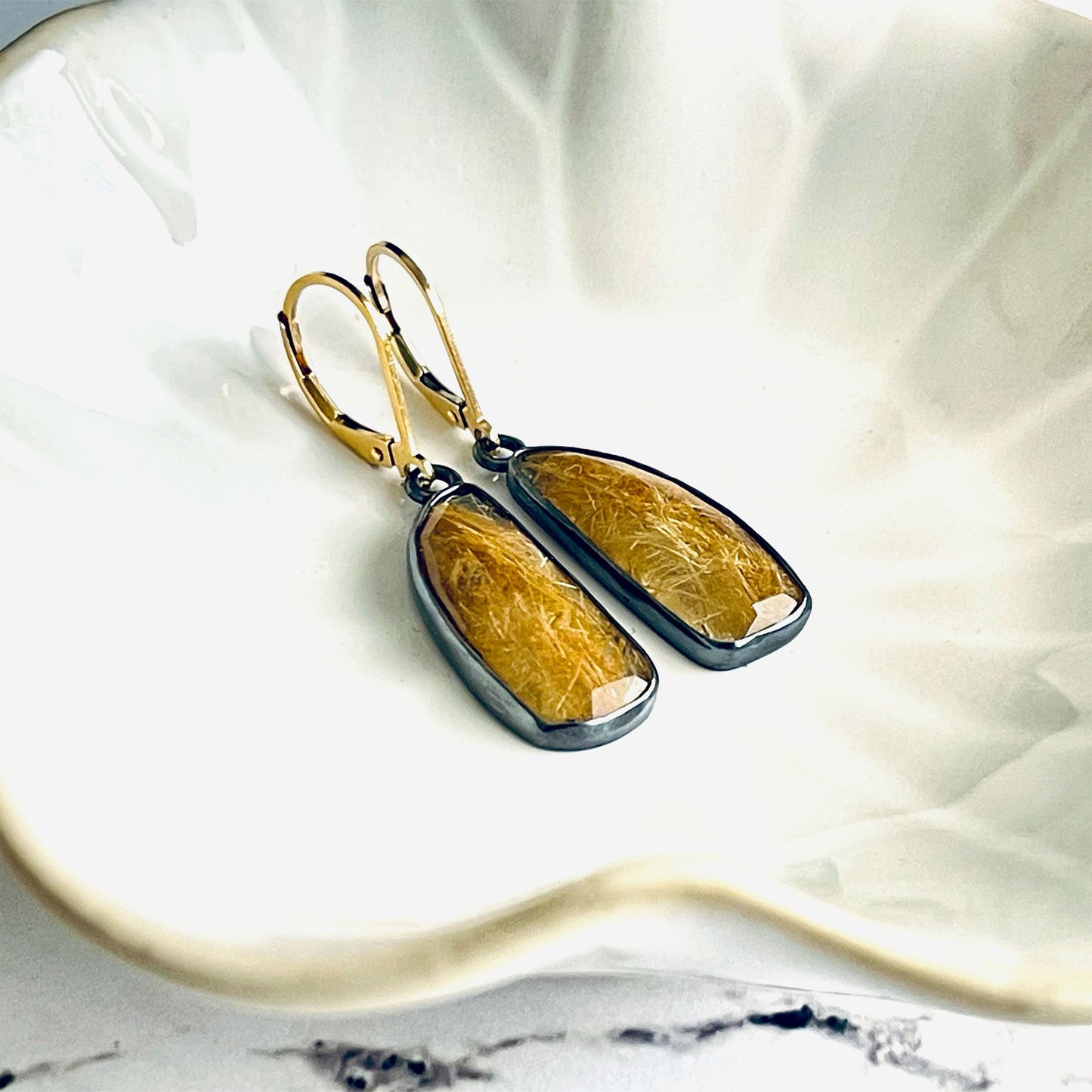 A pair of handmade sterling silver drop earrings featuring gold rutile quartz gemstones. The sterling silver bezel settings have been heavily oxidised and the earwires are 14k gold fill lever backs. The earrings are lying in a cream coloured dish. 