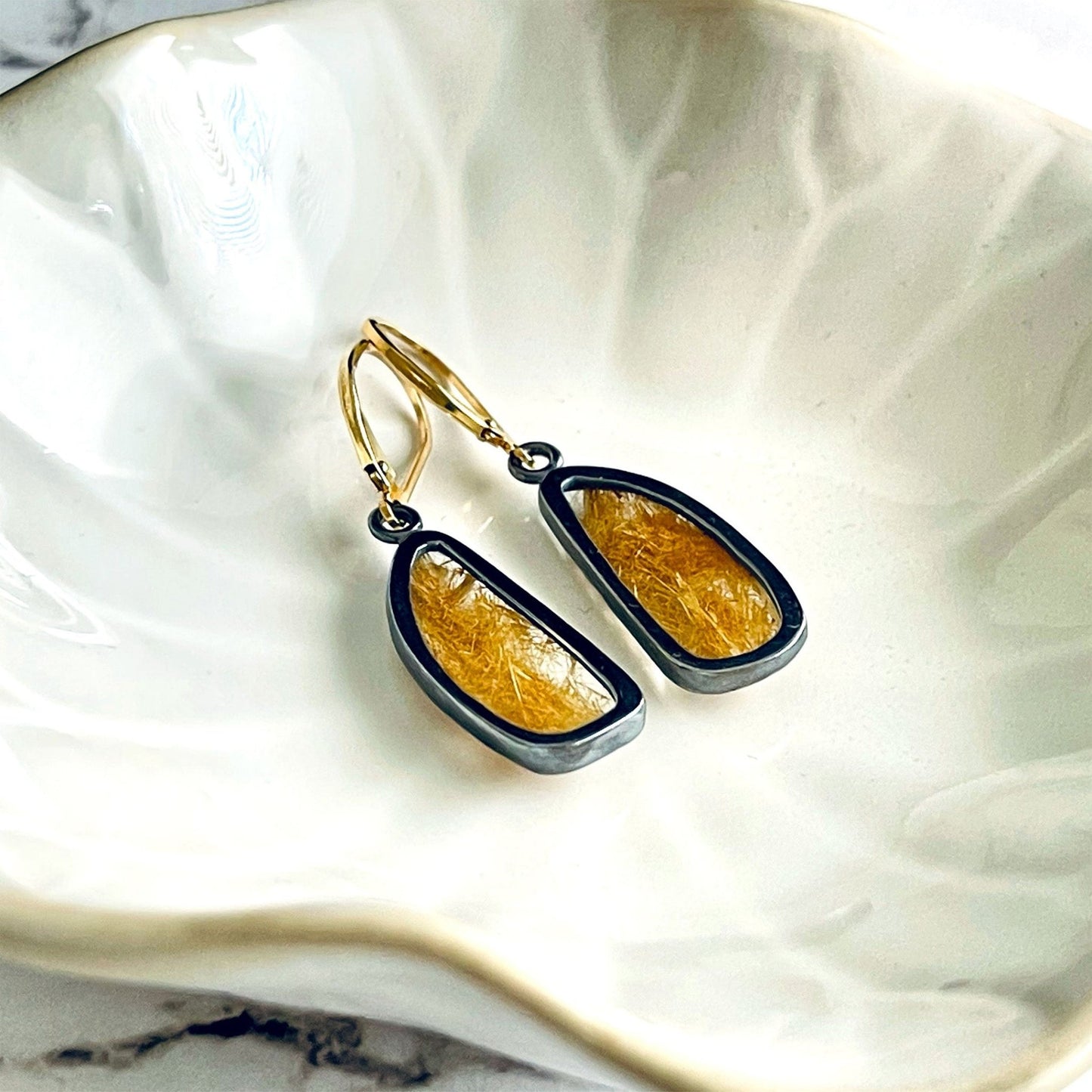 A pair of handmade sterling silver drop earrings featuring gold rutile quartz gemstones. The sterling silver bezel settings have been heavily oxidised and the earwires are 14k gold fill lever backs. The earrings are lying in a cream coloured dish. 