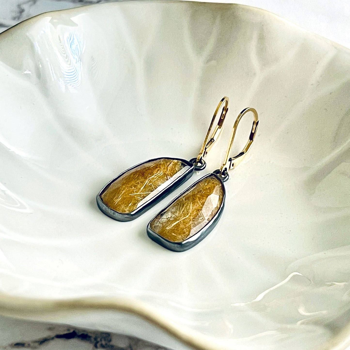 A pair of handmade sterling silver drop earrings featuring gold rutile quartz gemstones. The sterling silver bezel settings have been heavily oxidised and the earwires are 14k gold fill lever backs. The earrings are lying in a cream coloured dish. 