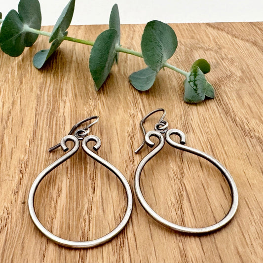 Oxidised sterling silver hoop earrings on a wooden surface with a small sprig of eucalyptus leaves.