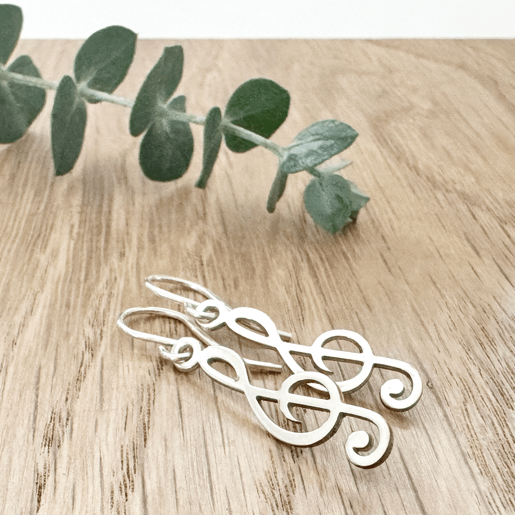 Sterling silver earrings with a treble clef design on a wooden surface with a small sprig of eucalyptus leaves.