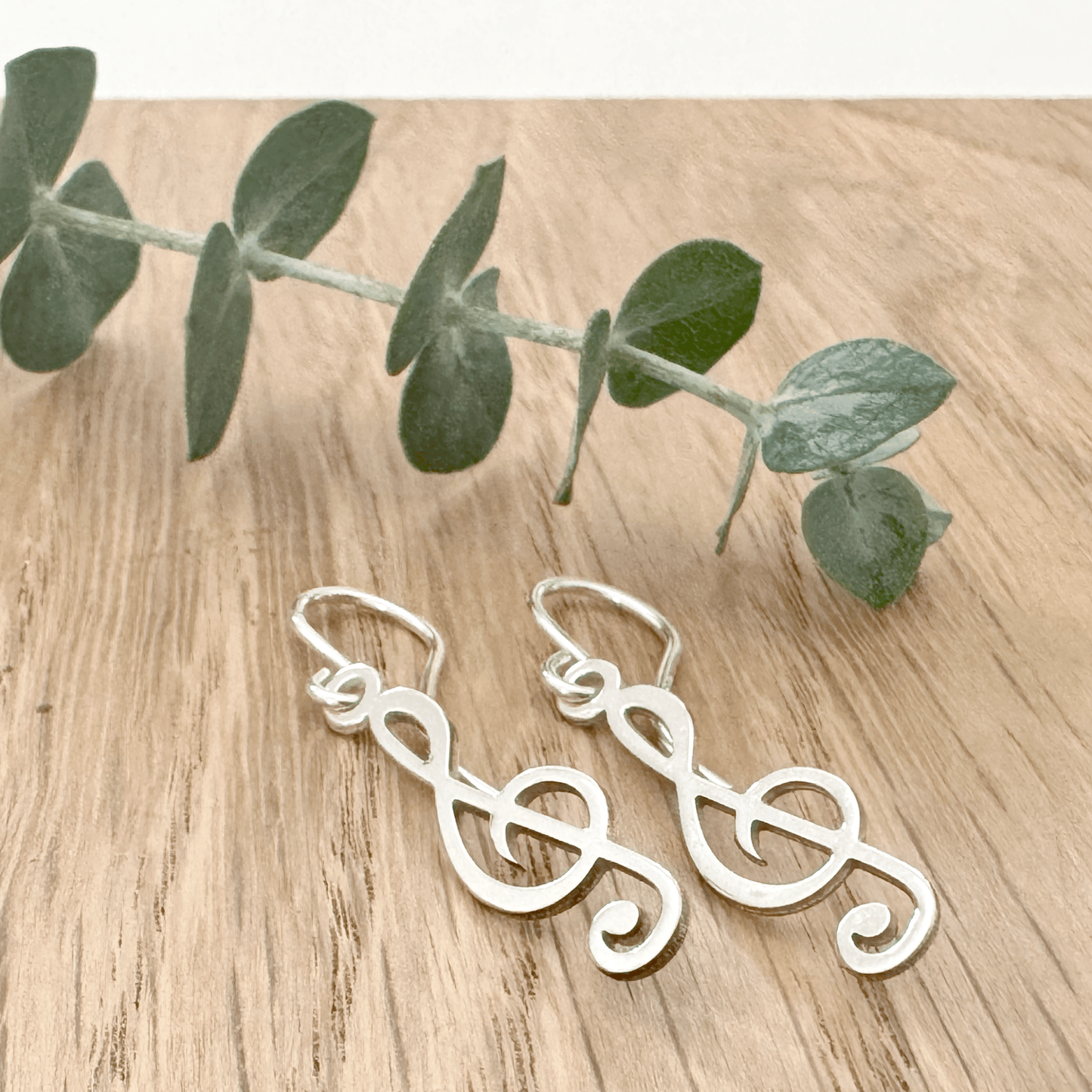 Sterling silver earrings with a treble clef design on a wooden surface with a small sprig of eucalyptus leaves.