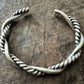 A sterling silver cuff bracelet on a wooden background. The cuff bracelet is comprised of two strands of silver, loosely twisted together. One is round and smooth, the other is very tightly twisted. It has been oxidised.