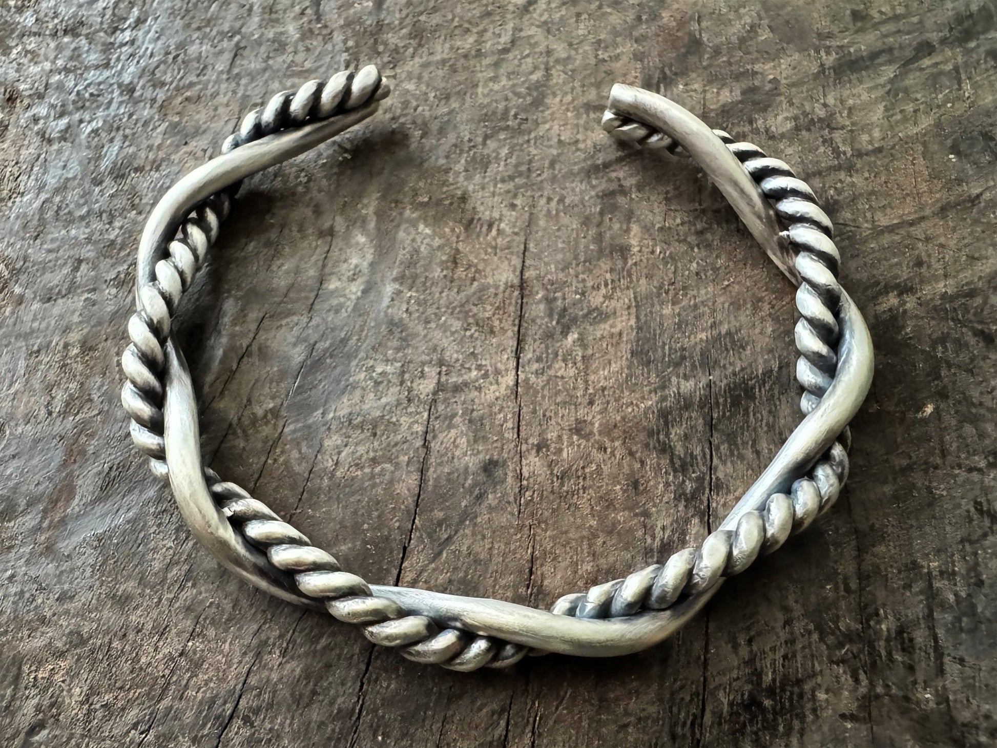 A sterling silver cuff bracelet on a wooden background. The cuff bracelet is comprised of two strands of silver, loosely twisted together. One is round and smooth, the other is very tightly twisted. It has been oxidised.