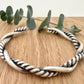 A sterling silver cuff bracelet on a wooden background with a small sprig of eucalyptus leaves. The cuff bracelet is comprised of two strands of silver, loosely twisted together. One is round and smooth, the other is very tightly twisted. It has been oxidised.