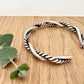 A sterling silver cuff bracelet on a wooden background with a small sprig of eucalyptus leaves. The cuff bracelet is comprised of two strands of silver, loosely twisted together. One is round and smooth, the other is very tightly twisted. It has been oxidised.