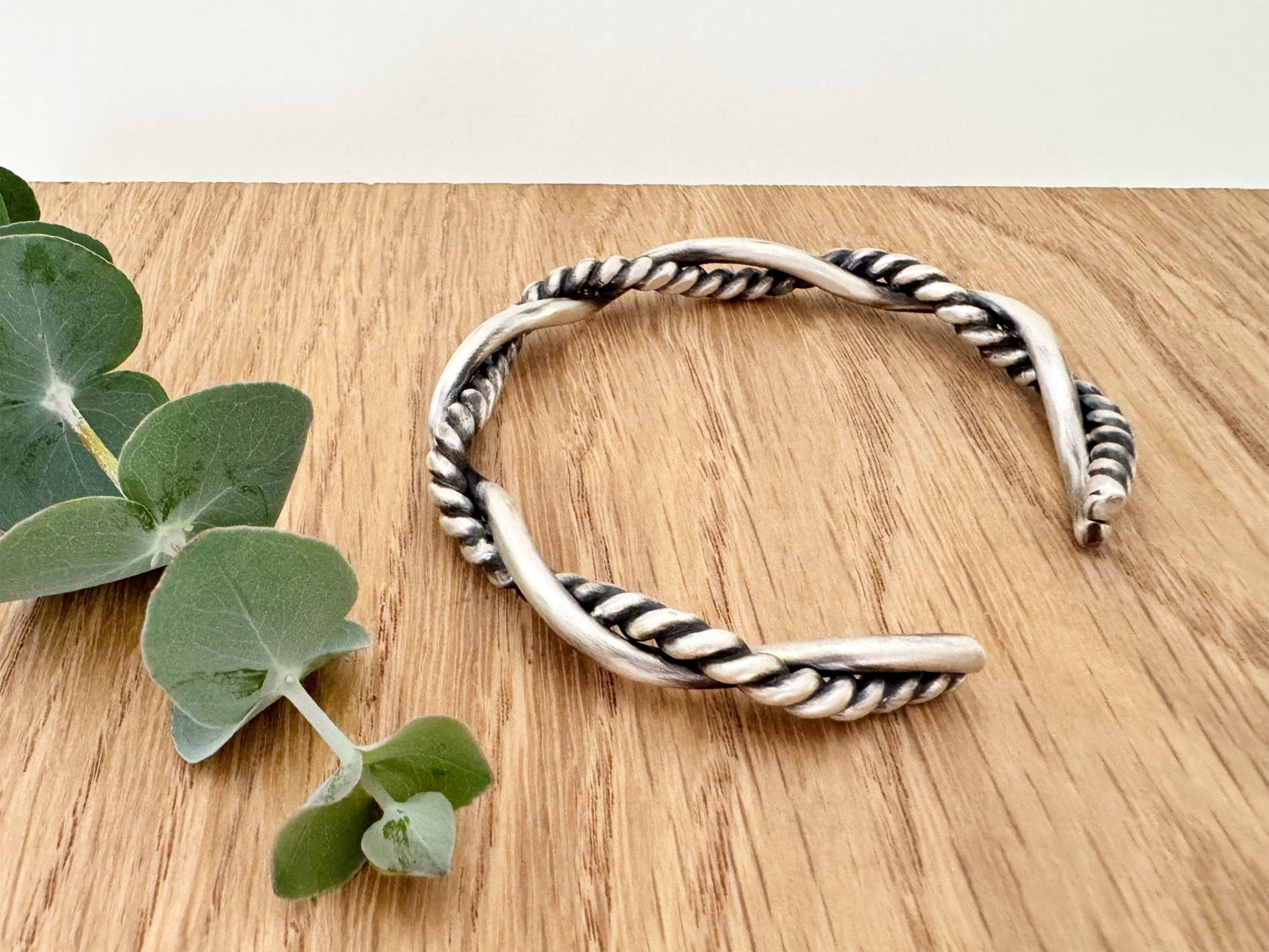 A sterling silver cuff bracelet on a wooden background with a small sprig of eucalyptus leaves. The cuff bracelet is comprised of two strands of silver, loosely twisted together. One is round and smooth, the other is very tightly twisted. It has been oxidised.