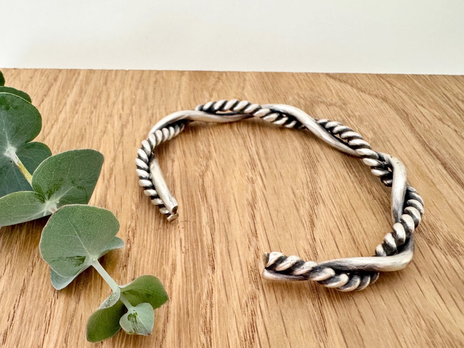 A sterling silver cuff bracelet on a wooden background with a small sprig of eucalyptus leaves. The cuff bracelet is comprised of two strands of silver, loosely twisted together. One is round and smooth, the other is very tightly twisted. It has been oxidised.