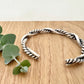A sterling silver cuff bracelet on a wooden background with a small sprig of eucalyptus leaves. The cuff bracelet is comprised of two strands of silver, loosely twisted together. One is round and smooth, the other is very tightly twisted. It has been oxidised.