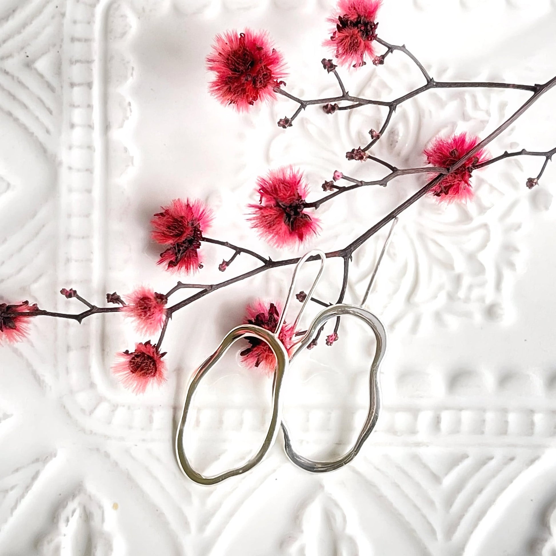 A pair of unique, abstract shaped sterling silver earrings, sitting on a textured white dish. There is dyed red blossom behind the earrings. Handmade by Australian jeweller Rebecca Cordingley.