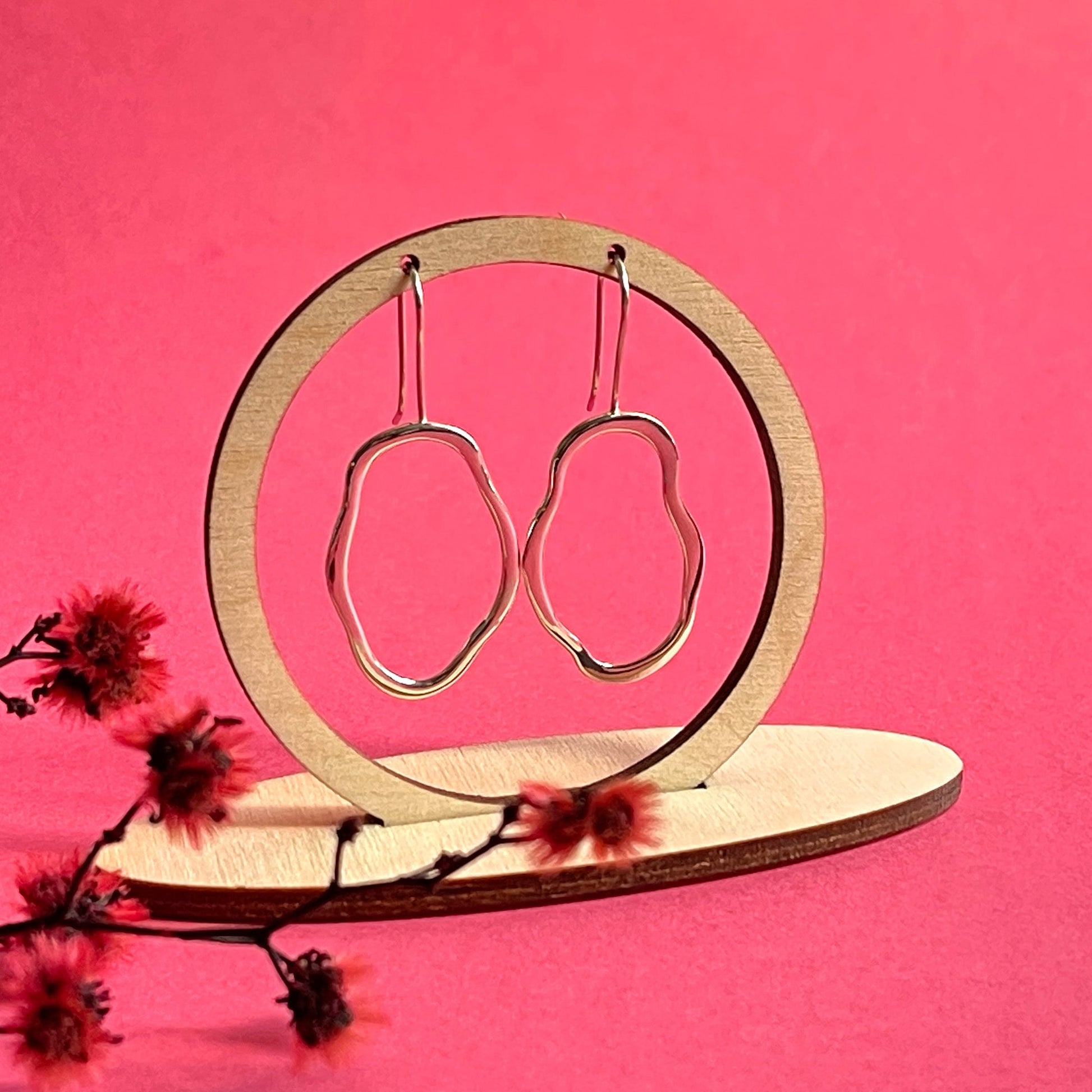 A pair of unique, abstract shaped sterling silver earrings, hanging from a wooden frame on a bright pink background. There is dyed red blossom in front of the frame. Handmade by Australian jeweller Rebecca Cordingley.