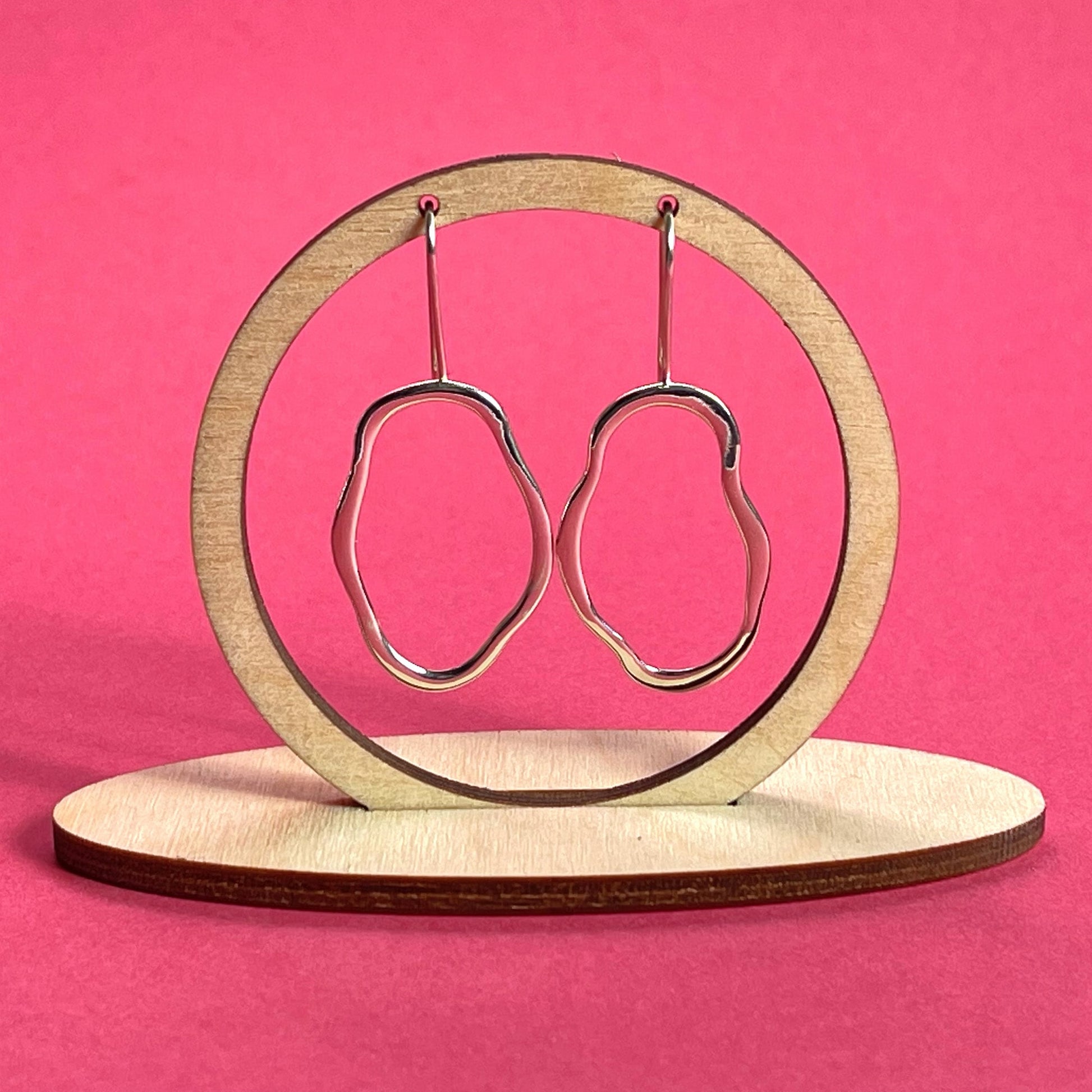 A pair of unique, abstract shaped sterling silver earrings, hanging from a wooden frame on a bright pink background. Handmade by Australian jeweller Rebecca Cordingley.