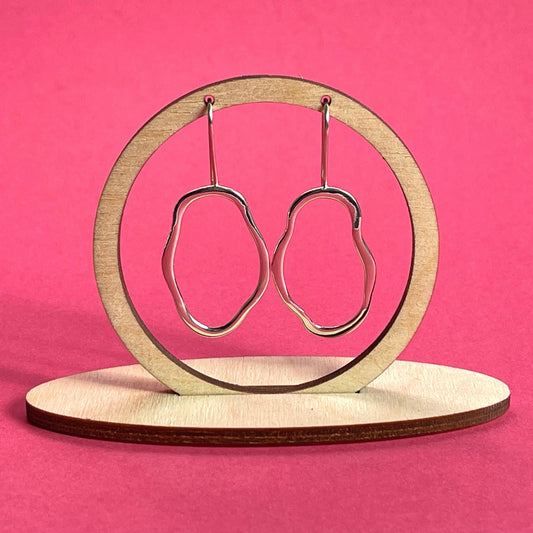 A pair of unique, abstract shaped sterling silver earrings, hanging from a wooden frame on a bright pink background. Handmade by Australian jeweller Rebecca Cordingley.