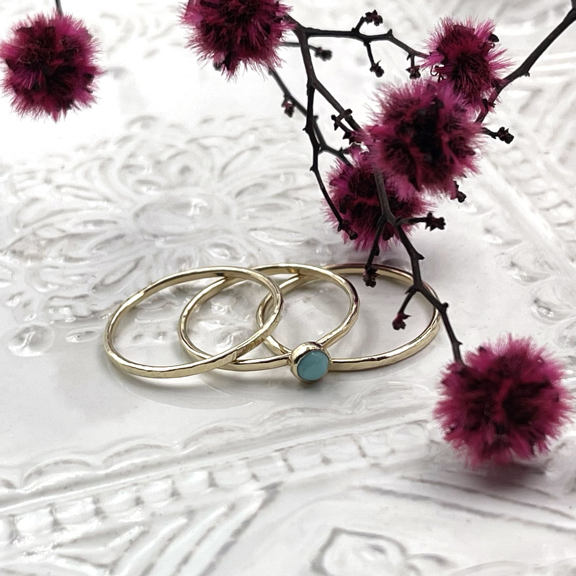Set of 3 dainty gold rings with a turquoise cabochon on a white textured dish with dried purple flowers