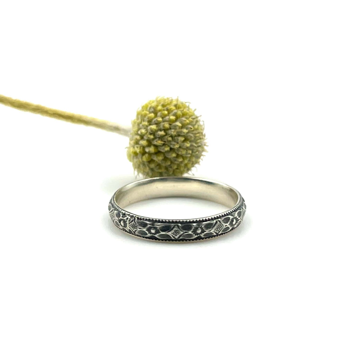 Simple silver ring with floral pattern in front of a dried billy button